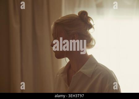 Jeune femme caucasienne adulte debout dans le profil près de la fenêtre, reflétant tranquillement dans le cadre de la maison, exprimant la pleine conscience calme et les soins de soi. Utile pour la santé mentale, le bien-être, les campagnes de style de vie Banque D'Images