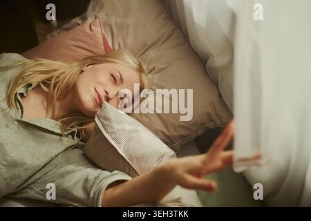 Jeune femme caucasienne adulte se reposant dans son lit, tendant vers le rideau et se réveillant à la maison. Représentant la routine matinale, le bien-être du sommeil, la publicité de style de vie, plan horizontal Banque D'Images