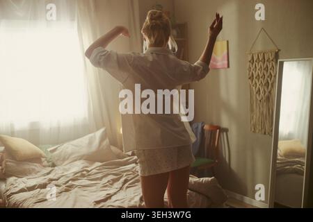 Jeune femme caucasienne adulte dansant à côté du lit après le réveil, commençant la routine du matin à la maison. Concept de soins personnels, bien-être, mode de vie sain, vue arrière moyenne Banque D'Images
