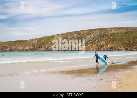 Deux surfeurs en combinaisons marchent le long du bord de l'eau à la Baie des Trépassés, portant leurs planches vers le surf. L'arrière-plan présente des rochers abrupts Banque D'Images