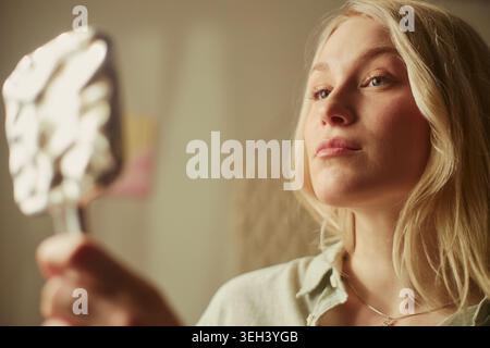 Portrait de jeune femme adulte caucasienne examinant le visage dans un miroir à main, exprimant l'auto-évaluation et la routine de beauté. Idéal pour les soins de la peau, les cosmétiques, le bien-être, le marketing de soins personnels Banque D'Images
