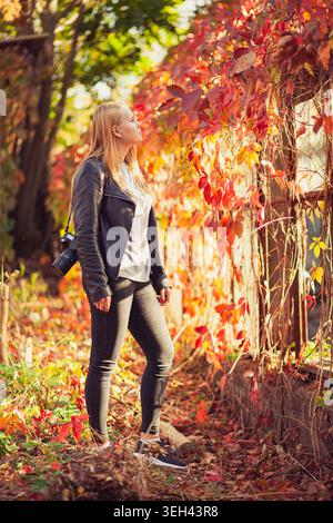 Jeune femme blonde avec caméra explore le parc d'automne. Elle regarde des feuilles rouges vibrantes dans la lumière chaude du soleil. La photo Lifestyle capture la créativité, le passe-temps et Banque D'Images