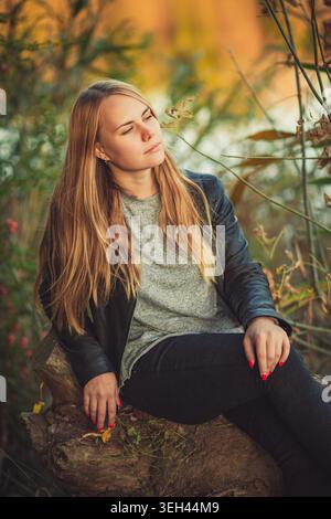 Jeune femme avec de longs cheveux blonds est assise sur la bûche dans la forêt d'automne. Elle porte des vêtements décontractés et a l'air réfléchie. Lumière naturelle douce et couleurs chaudes créa Banque D'Images