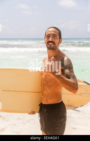 Portrait en gros plan d'un surfeur souriant avec des tatouages et un chignon d'homme faisant un geste shaka tout en tenant une planche de surf jaune vintage. Banque D'Images