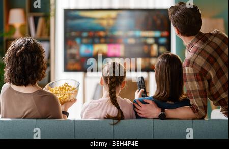 Une famille heureuse profite de moments de qualité ensemble dans un salon moderne, regardant le contenu de la télévision en streaming sur une grande télévision à écran plat. L'atmosphère i Banque D'Images