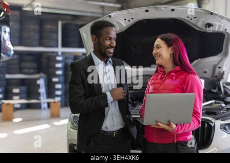 Mécanicien féminin utilisant un ordinateur portable pour le diagnostic tout en parlant avec un client dans un atelier avec des voitures sur cric et de l'équipement professionnel. Banque D'Images