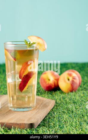 Vue détaillée d'un grand verre rempli de thé glacé à la pêche froide, de menthe fraîche et de tranches de fruits juteux sur une pelouse verte Banque D'Images