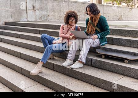 Diverses étudiantes étudient ensemble sur les marches du campus, partageant ordinateur portable avec skateboard à proximité Banque D'Images