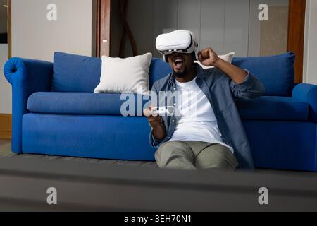 Homme afro-américain dans sa vingtaine jouant au jeu VR devant le canapé bleu, tenant le contrôleur Banque D'Images