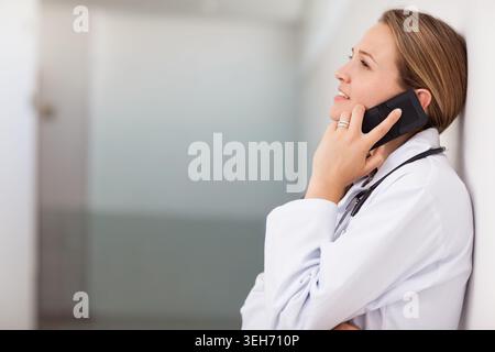 Médecin féminin appuyé contre le mur dans le couloir tenant un téléphone noir portant un manteau blanc, espace de copie Banque D'Images