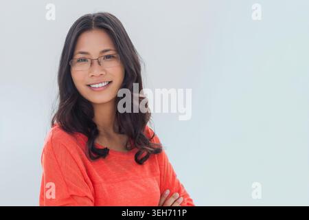 Femme asiatique debout face à la caméra en studio, portant un haut à manches longues orange et des lunettes transparentes Banque D'Images