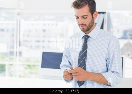 Homme portant chemise à carreaux, cravate, tenant le téléphone, regardant le moniteur dans le bureau avec des fenêtres, espace de copie Banque D'Images