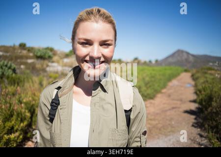 Femme debout, souriante sur un sentier de randonnée ensoleillé portant une chemise olive et un sac à dos avec des sangles bronzées Banque D'Images