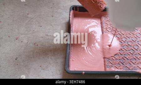 Gros plan du rouleau de peinture dans le bac avec de la peinture rose. Le fond de plancher de béton a des éclaboussures, suggérant la rénovation ou la décoration de maison de bricolage. Concept de Re Banque D'Images