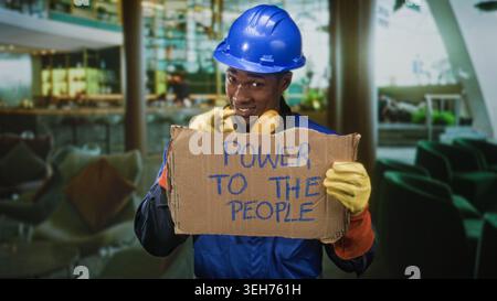 Homme ouvrier de la construction tenant le panneau en carton lisant le pouvoir aux gens, portant un casque, des protections auditives et des gants tout en souriant légèrement dans un bâtiment Banque D'Images