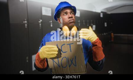 Homme constructeur tenant un panneau en carton lisant agir maintenant et faisant des gestes vers le haut dans un vestiaire d'un bâtiment tout en portant un casque et des gants ; plaidoyer urgent. Banque D'Images
