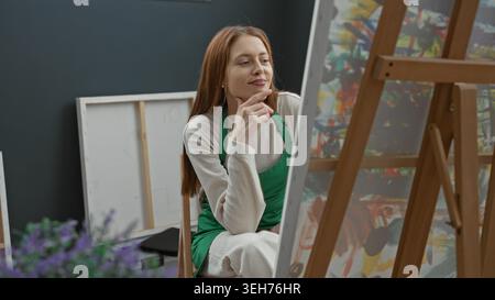 Femme peintre dans le tablier vert touche le menton tout en étant assis à chevalet en bois avec toile et peintures dans l'atelier d'art ; contemplation calme focus. Banque D'Images