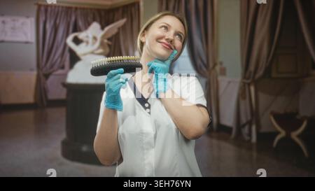 Femme dentiste en uniforme blanc tient le guide de teinte de dent et touche la joue avec la main gantée dans la construction de la clinique ; soins dentaires de confiance. Banque D'Images