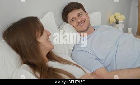 Femme souriant sur le lit avec la main sur l'oreiller tandis que l'homme touche sa taille, anneaux visibles, jeune couple détendu appuyé sur des oreillers blancs dans un bâtiment ; inti Banque D'Images