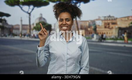 Femme aux cheveux bouclés pointe le doigt vers le haut et sourit largement tout en se tenant debout sur une rue de la ville bordée de bâtiments historiques et de piétons ; curiosité de joie Banque D'Images