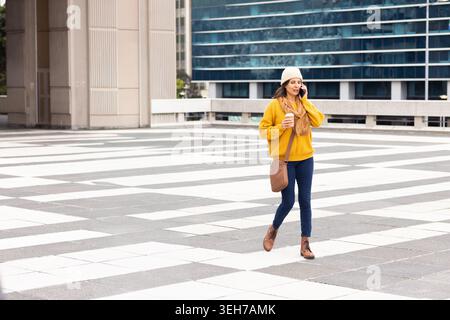 Femme mature marchant à travers la place à motifs dans le pull moutarde tenant le café et le téléphone, espace de copie Banque D'Images