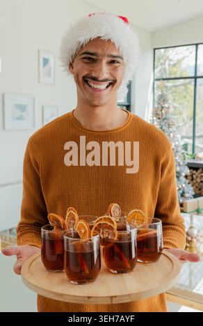 Homme portant chandail moutarde chapeau de Père Noël tenant une planche de bois ronde avec six verres et orange séchée Banque D'Images