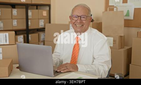Homme souriant avec les mains sur l'ordinateur portable tapant au bureau parmi les colis empilés et les boîtes dans un bâtiment de salle d'expédition ; confiance entrepreneuriale. Banque D'Images