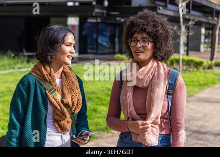 Diverses amies féminines marchant et discutant le long du chemin du campus, tenant le téléphone et portant des sacs à dos. Duo, camarades de classe, à l'extérieur, université, passerelle, conversation, smartphone Banque D'Images