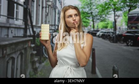 Femme tient la barre de chocolat blanc dans une main tout en touchant ses cheveux et en se bourrant les lèvres dans une rue ; décision de tentation. Banque D'Images