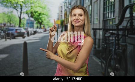 Femme pointe le doigt vers le haut avec la main droite et pointe vers la droite avec la main gauche tout en souriant dans la rue ; invitation de confiance. Banque D'Images
