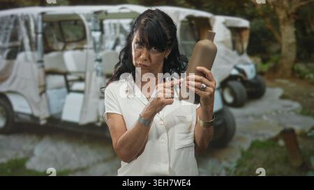 Femme âgée en chemisier blanc pointe doigt à la bouteille de poterie tout en inspectant l'étiquette près des chariots de golf garés dans la forêt ; préoccupation. Banque D'Images