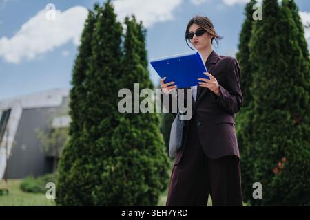 Femme d'affaires en costume sombre examinant des documents à l'extérieur avec presse-papiers bleu près des arbres à feuilles persistantes Banque D'Images