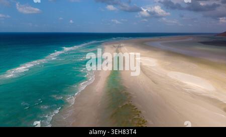 Vue aérienne d'un long banc de sable de sable rencontrant l'océan turquoise avec des vagues douces, lagon peu profond sur la droite, et ciel nuageux spectaculaire ; tropical isolé c Banque D'Images