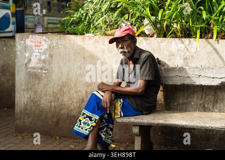 Sri Lanka - 20 février 2026 : vue d'un homme âgé avec une barbe grise et un chapeau rouge assis sur un banc de pierre portant un motif traditionnel bleu saron Banque D'Images