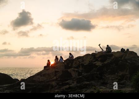 Sri Lanka - 25 January 2026: View of people sitting on a rocky cliff overlooking the ocean during a golden sunset with soft clouds. Banque D'Images
