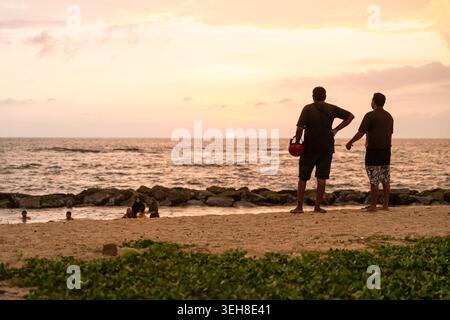 Sri Lanka - 14 janvier 2026 : vue de deux hommes debout sur une plage de sable fin regardant le coucher du soleil sur l'océan Indien avec des gens nageant près du rivage. Banque D'Images
