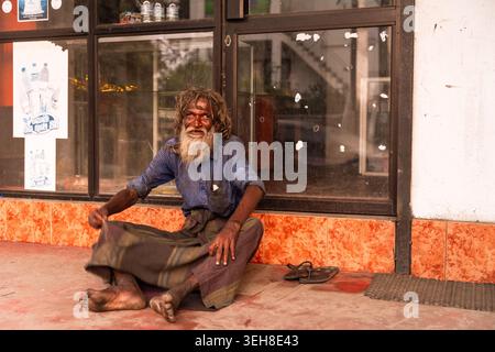 Sri Lanka - 14 janvier 2026 : vue d'un homme âgé avec une longue barbe et des cheveux désordonnés assis par terre devant une vitrine de magasin avec des affiches. Banque D'Images