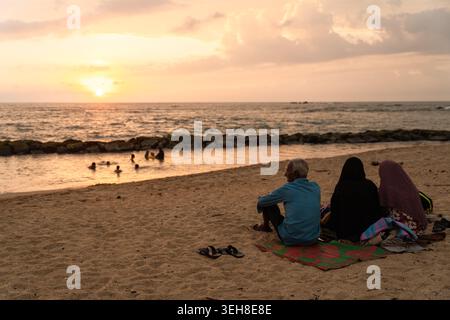 Sri Lanka - 14 janvier 2026 : vue de personnes assises sur une plage de sable et d'autres nageant dans l'océan pendant un coucher de soleil doré au ciel nuageux. Banque D'Images