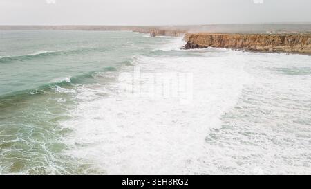 Vue aérienne des falaises escarpées de Sagres avec de puissantes vagues blanches s'écrasant contre la côte rocheuse sous un ciel brumeux à Sagres, Faro, Portugal. Banque D'Images