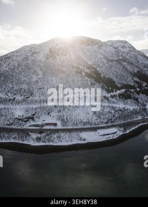 Vue aérienne du Grotfjord avec un sommet de montagne enneigé, une route côtière et le soleil brille sur la crête à Tromvik, Troms, Norvège. Banque D'Images