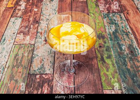 Une tasse en verre contient une mousse de mangue jaune crémeuse surmontée de tranches de mangue fraîches, reposant sur une surface en bois rustique multicolore. Banque D'Images
