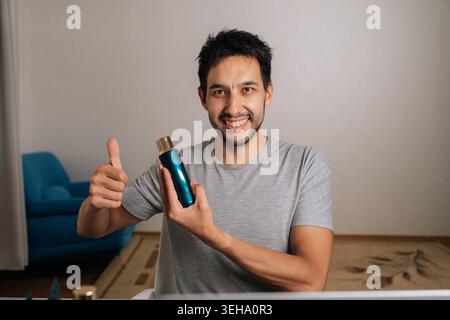 Vlogger masculin positif donnant les pouces vers le haut regardant la caméra souriante et tenant le produit cosmétique pendant un appel vidéo en ligne, examinant les articles de beauté pour les réseaux sociaux Banque D'Images