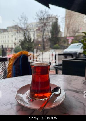 Thé turc traditionnel dans le verre en forme de tulipe se dresse sur la table du café près de la fenêtre de la rue pluvieuse avec un fond urbain flou, une atmosphère chaleureuse Banque D'Images