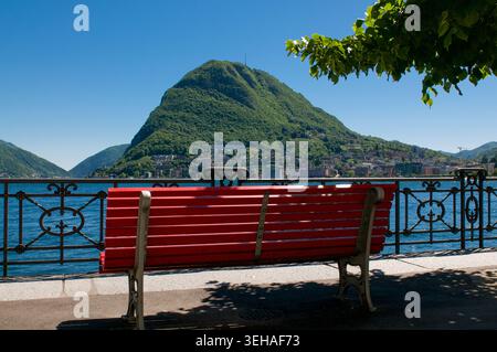 Vue panoramique panoramique d'un banc rouge classique vide sur la promenade au bord du lac de Lugano, Tessin, Suisse. Magnifique paysage alpin avec mou Banque D'Images