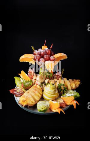 Élégant arrangement de fruits avec verre de martini de raisins, ananas sculpté, kiwi et tranches d'orange sur fond sombre Banque D'Images