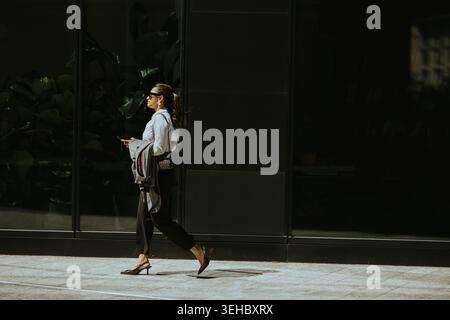 Une femme marche le long d'un trottoir de la ville, tenant un téléphone et portant des vêtements décontractés. Elle passe devant un grand bâtiment en verre. Le soleil brille, et l'ombre Banque D'Images