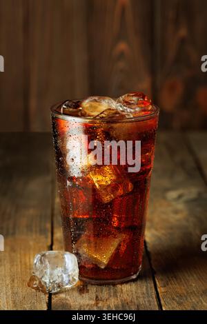 Rafraîchissant cola pétillant avec glaçons fondants dans un verre créant de la condensation sur fond de bois rustique Banque D'Images