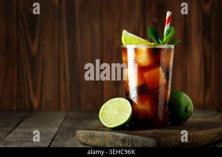 Cuba libre Drink, avec du cola, du rhum, des glaçons, du citron vert et des feuilles de menthe fraîche, debout sur une surface en bois avec un fond rustique Banque D'Images