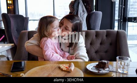 Mère embrassant sa fille au café, scène de petit déjeuner confortable avec tranche de pizza et gâteau au chocolat sur la table en bois, mère tenant l'enfant sur le tour Banque D'Images