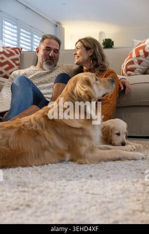 Couple assis sur un canapé léger à la maison, femme penchée, grand Golden retriever couché, chiot curling. Confortable, chiens, affection, salon, tapis, coussins, plante Banque D'Images